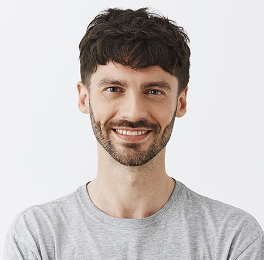 Man with short curly dark hair and beard in a light gray t-shirt, smiling against white background