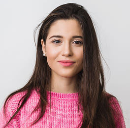 Woman with long brown hair in a bright pink sweater, smiling slightly against white background